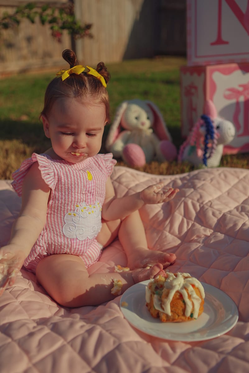 A baby sitting on a pink quilt with a bundt cake