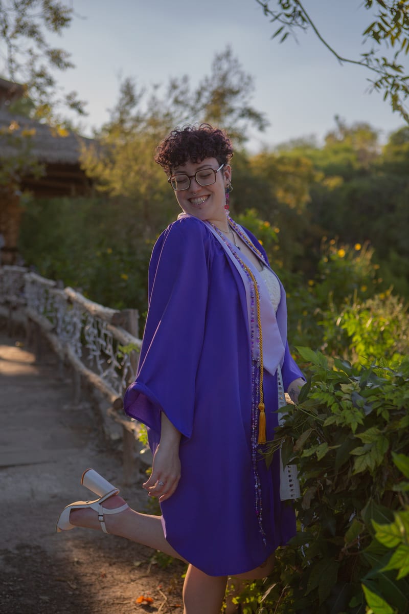 A graduate in a purple gown kicking up one heel on a garden path, from a graduation portrait session