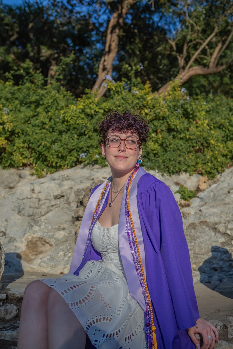 A graduate in a purple gown seated on limestone rocks, from a graduation portrait session