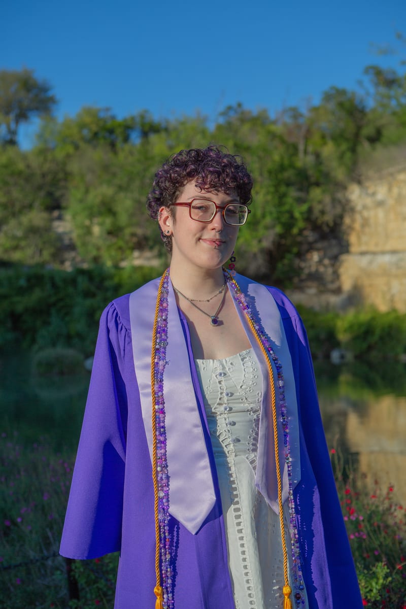 A graduate in a purple gown standing by the water, from a graduation portrait session